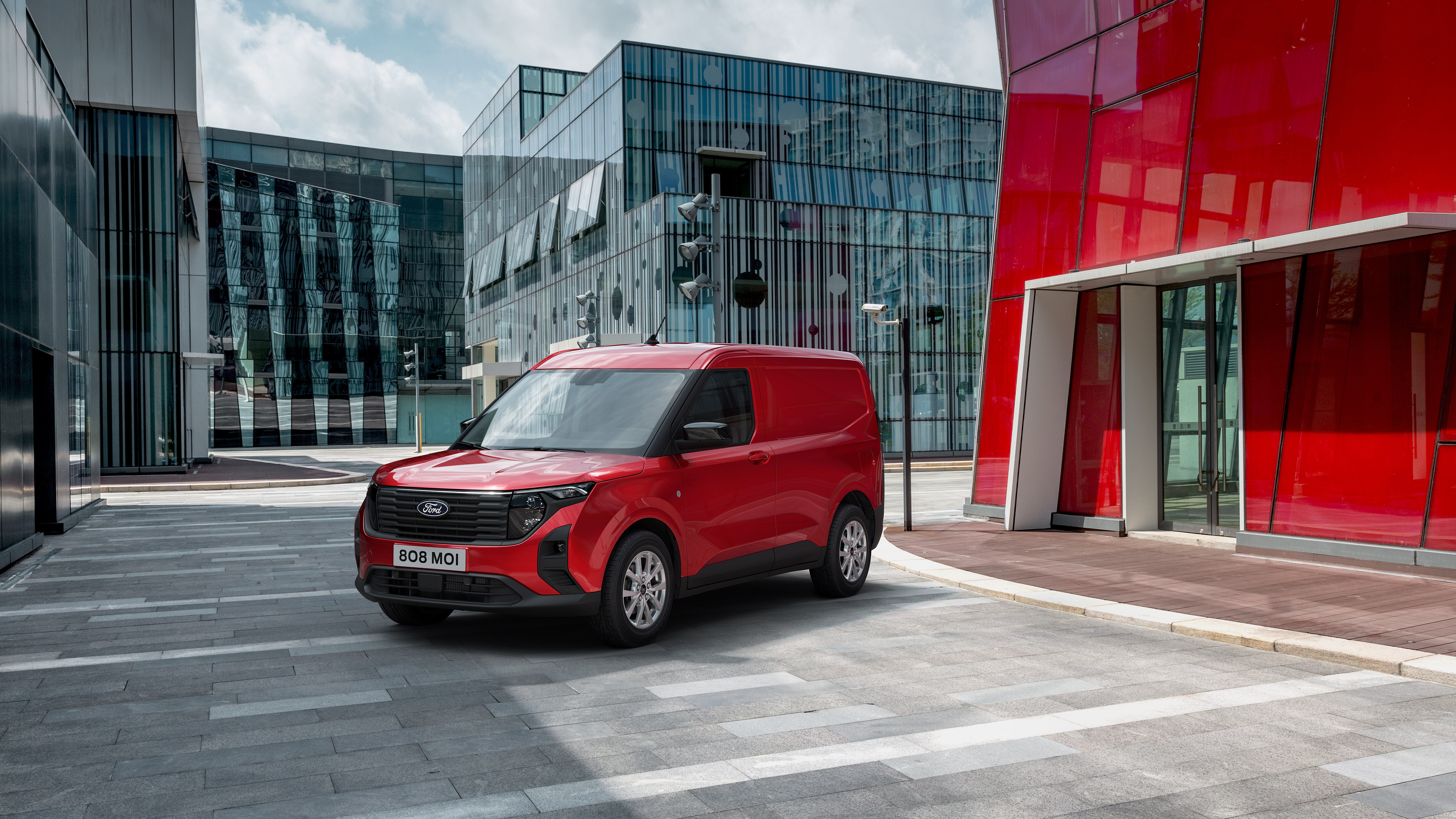 A red compact van parked in a modern corporate square, sunny